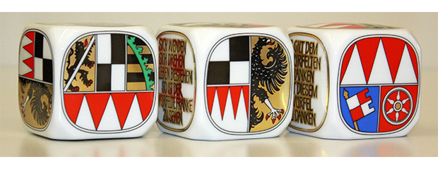 Drei Würfel aus weißem Porzellan liegen auf einem beige-farbenem Tisch. Auf den Würfeln sind die Wappen der drei fränkischen Regierungsbezirke aufgemalt.