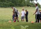 Fünf männliche Jugendliche in Sportklamotten stehen auf einem grünen Golfplatz. Zwei Teenager halten Golfschläger in der Hand, einer spielt damit gerade einen Ball, der andere schaut einem Ball hinterher.