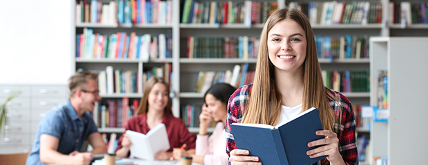 Symbolfoto: Junge Frau in einer Bibliothek hält ein blaues Buch hoch und lächelt dabei